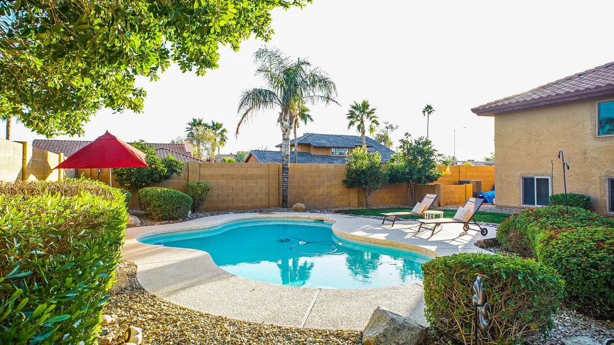 A vibrant backyard setting with a sparkling swimming pool, lounge chairs shaded by a red canopy, palm trees, and beautifully manicured bushes. The area is ideal for relaxing, sunbathing, or gathering with friends after a round of golf.