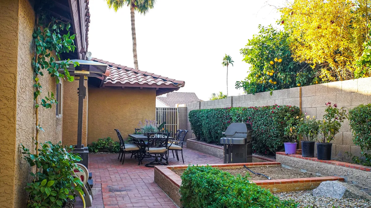 An outdoor dining set sits beneath a shaded patio, adjacent to a built-in grill, with palm trees and flourishing gardens in the background. The setup encourages gatherings for outdoor meals, barbecues, and celebrations, all within the lush greenery of the backyard.