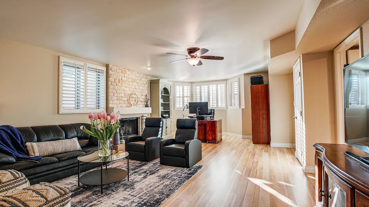A welcoming living area with a black leather sofa, coffee table adorned with flowers, a dark armchair, a decorative stone wall, and a visible home office setup in the corner. The space is multifunctional and stylish.
