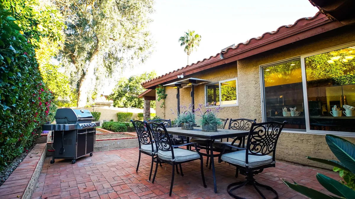 A cheerful backyard dining setup featuring a table with chairs, a barbecue grill, and lush greenery. The surrounding landscaping and relaxed ambiance suggest a perfect setting for gatherings, outdoor meals, and casual celebrations.