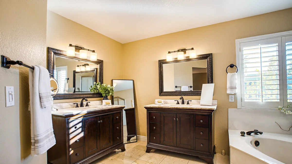 A spacious bathroom with dual vanities, large mirrors, a bathtub, and generous natural light from a nearby window. The décor is modern and spa-like, conveying a sense of relaxation and indulgence.