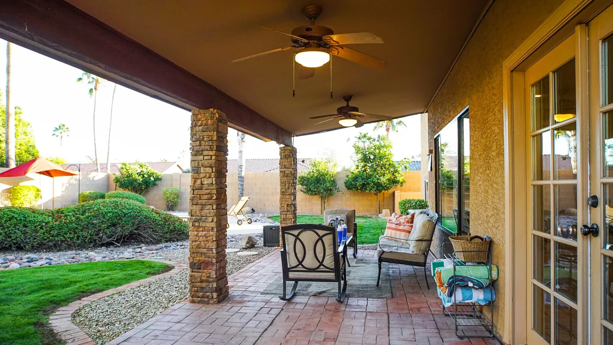 The photo shows a covered patio with stone columns, comfortable outdoor seating, decorative pillows, and a green lawn stretching beyond. The space is ideal for unwinding, entertaining, or having a casual business call in the fresh air.