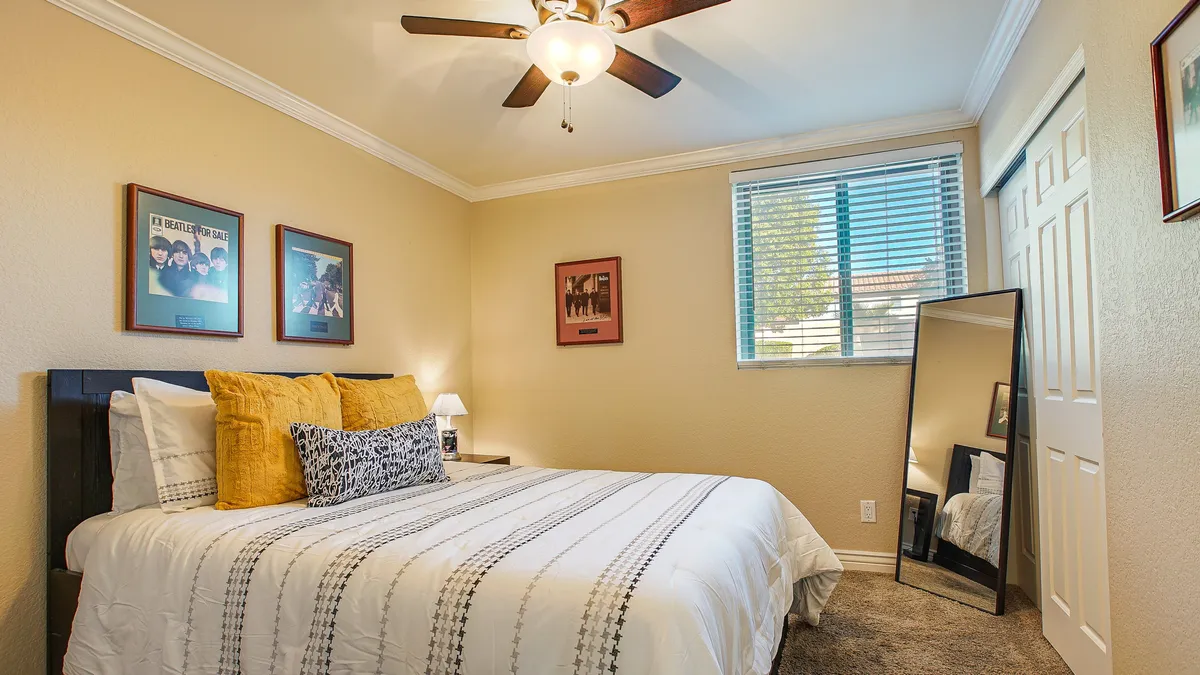 A cozy bedroom with a neatly made bed adorned with decorative pillows, framed pictures on the wall, a mirror, and a window with blinds letting in natural light. The room feels warm and inviting, perfect for relaxing or sleeping.