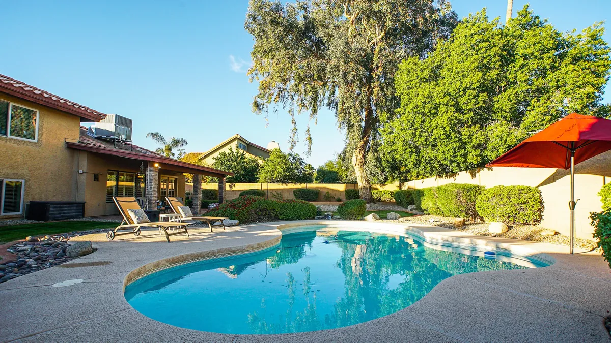 A sparkling swimming pool framed by inviting lounge chairs, a mature shade tree, and a striking red umbrella. The scene evokes a tranquil, resort-like atmosphere that highlights spaces for swimming, sunbathing, and relaxing by the water.