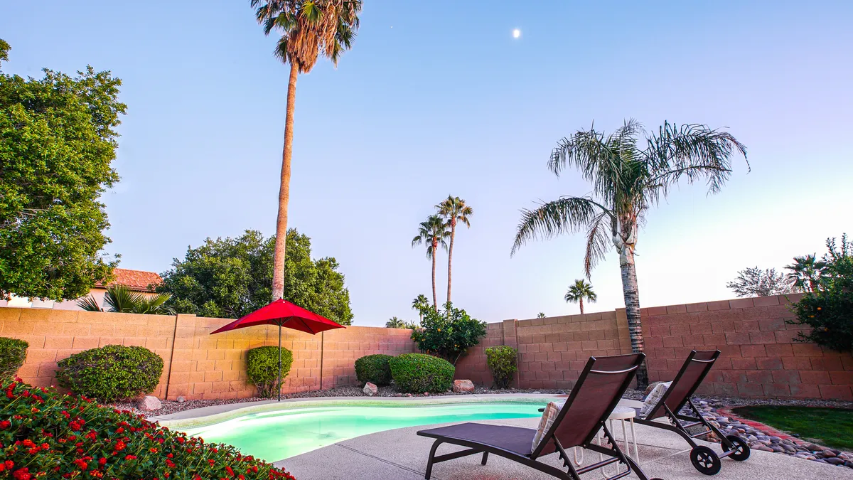A sparkling swimming pool is surrounded by lounge chairs, palm trees, and a bold red umbrella, with vibrant flower beds completing the inviting setting. The tranquil atmosphere invites relaxation for both owners and their pets.
