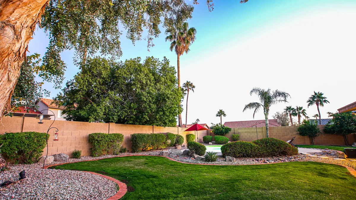 A wide stretch of lush green lawn is dotted with palm trees and anchored by a red patio umbrella, offering both sun and shade. The space is open and versatile, ideal for playing games like cornhole, relaxing, or letting children and pets play freely.