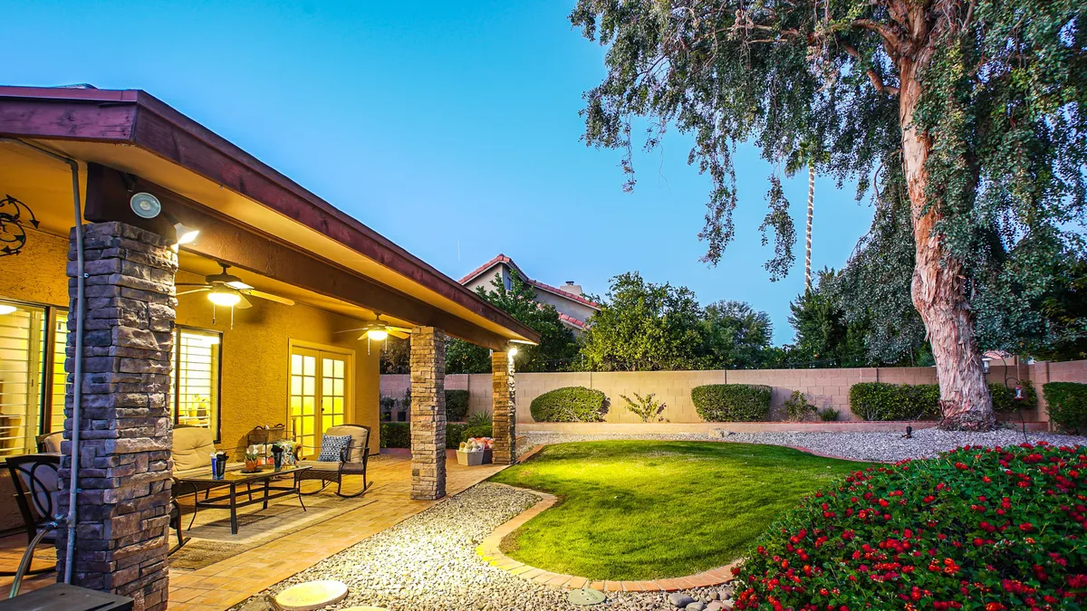 A covered patio with a dining area overlooks a lush green lawn and vibrant garden flowers beneath a canopy of mature trees. The scene captures opportunities for outdoor dining, gatherings, and tranquil relaxation.