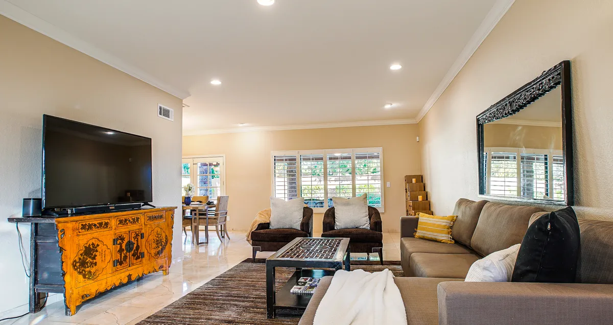 A spacious, inviting living room with a comfortable sofa, decorative pillows, a large television, and an ornate wooden cabinet. The dining area is visible in the background, showcasing the open-concept layout and natural flow between spaces.