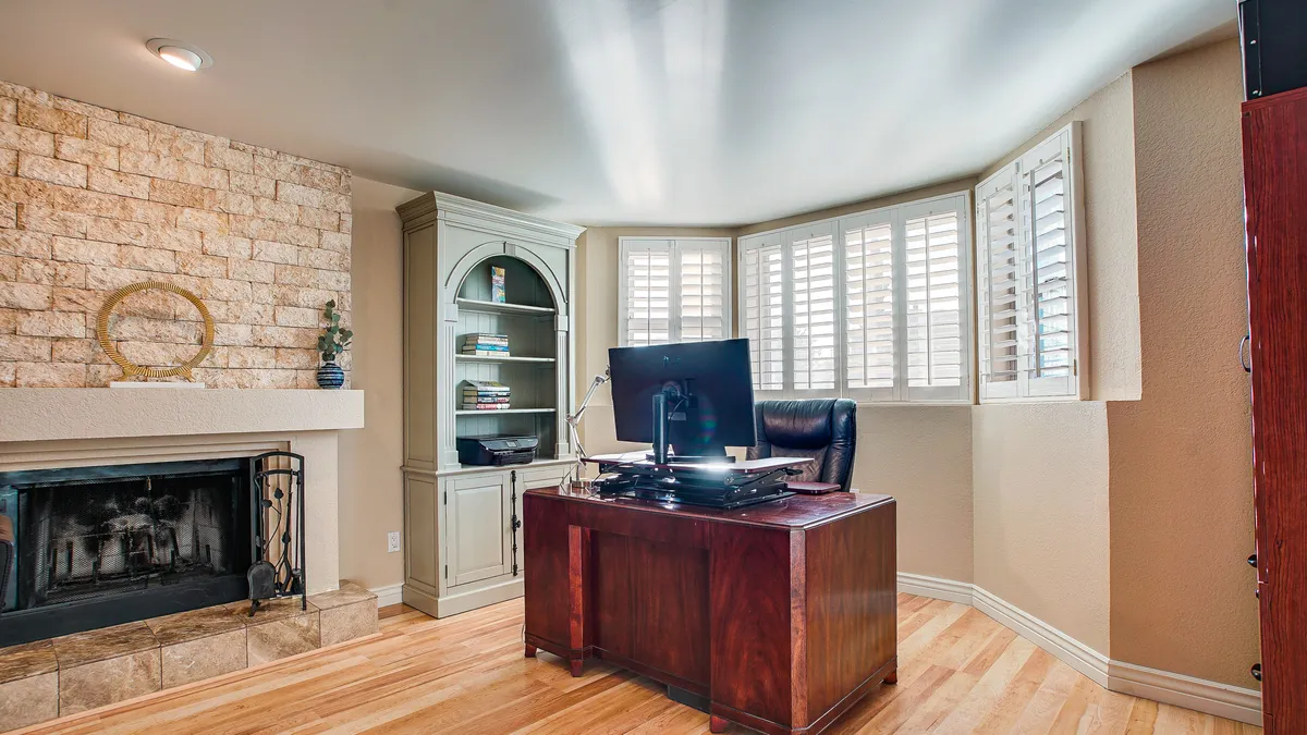 A serene home office space featuring a wooden desk set near a window with shutters, a bookshelf filled with books, and a fireplace adding warmth and coziness. The scene is organized and uncluttered, ideal for focused work or study sessions.